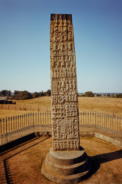 A standing stone in a field, covered in carvings of people