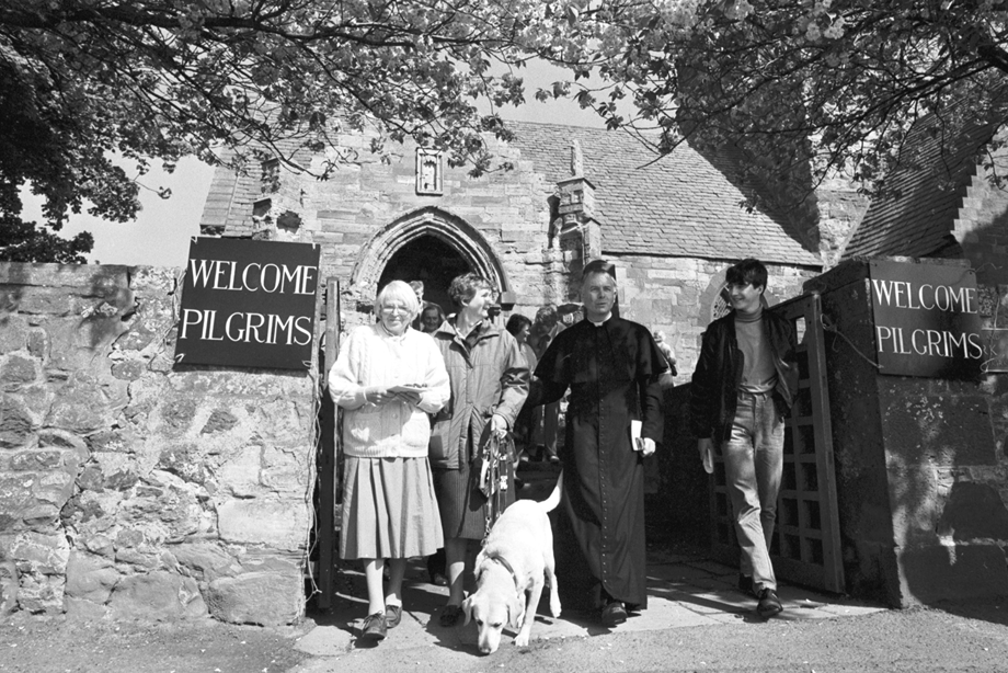 A group of people walking out of a church, which has 'welcome pilgrims' posters on the gates. Image reference number: 000-000-613-904-R 0