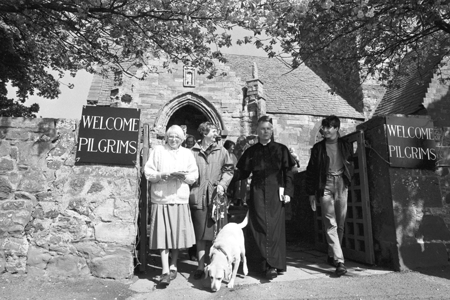 A group of people walking out of a church, which has 'welcome pilgrims' posters on the gates. Image reference number: 000-000-613-904-R 0
