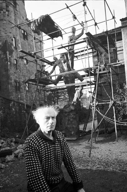 A person standing outside a stone building, and wooden sculpture surrounded by scaffolding. Reference no: 000-000-532-049-R