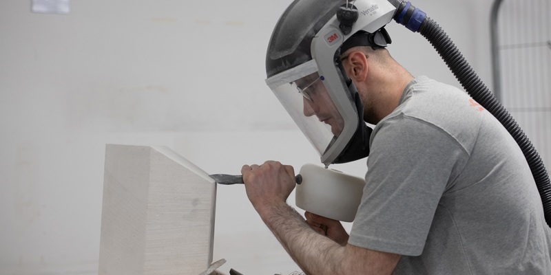 A stonemason wearing protective equipment works on a block of white stone