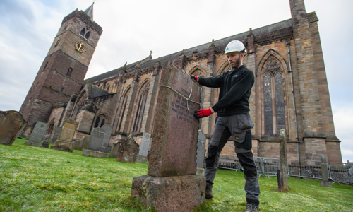Man in white Historic Environment Scotland helmet stands with two hands on a old gravestone. A piece of string is tied around the gravestone. Behind the man is Dunblane Cathedral, and we see the steeple on the left hand side.
