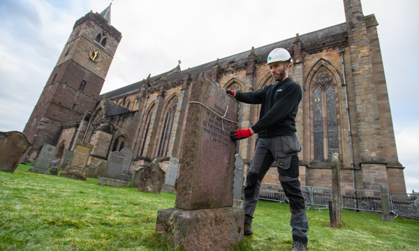 Man in white Historic Environment Scotland helmet stands with two hands on a old gravestone. A piece of string is tied around the gravestone. Behind the man is Dunblane Cathedral, and we see the steeple on the left hand side.