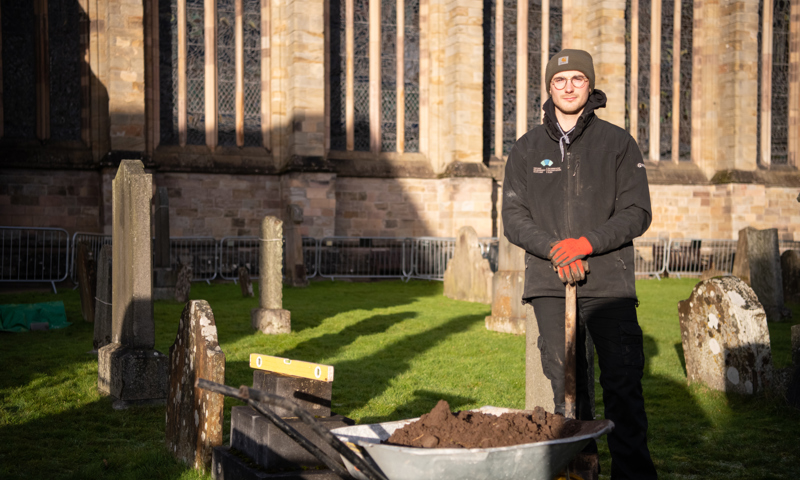 HES staff member with glasses and a beanie hat stands behind a wheelbarrow filled with dirt. The man is in a graveyard and a church wall is seen in the background.