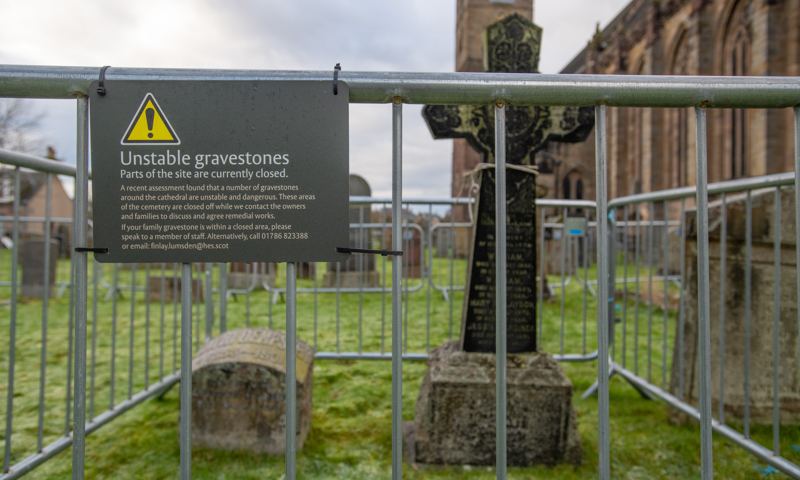 A sign hangs on a fence that surround two grave memorials. Part of the sign reads: "Unstable gravestones. Parts of the site are currently closed."