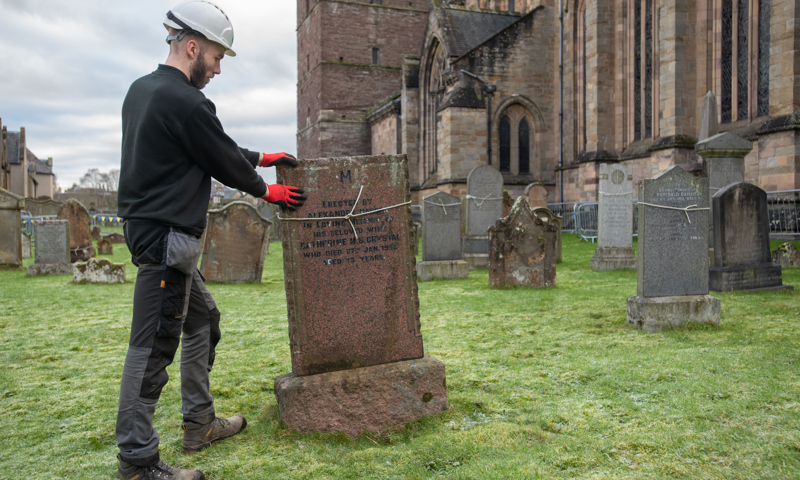 Man in white hat stands with two hands on a brown gravestone. The gravestone is listing to the left and has a piece of thin rope tied around it.