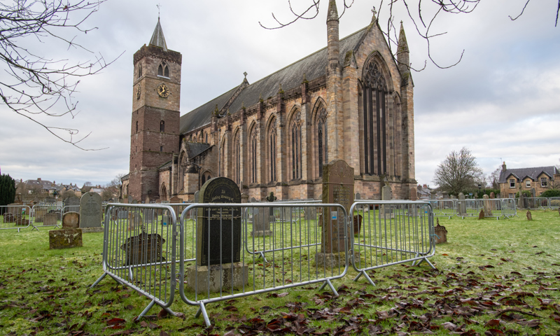 It is an overcast winter's day. The ground is wet and brown leaves are scattered on the grass around fencing that is in place surrounding various gravestones in a graveyard. In the background we see Dunblane Cathedral.