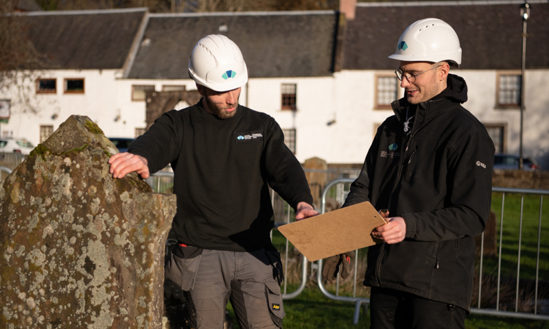 Two Historic Scotland inspectors in white hard hats inspect a gravestone. One is holding a clipboard, which the other is pointing at.
