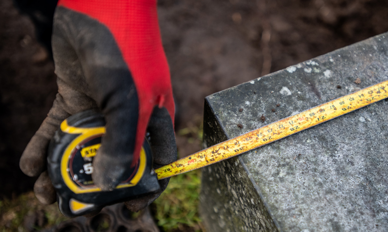 A gloved hand holds a measuring tape to the corner of a large, cut stone.