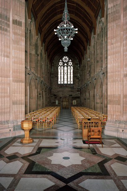 A stone church corridor, looking towards a large stained glass window with ornately carved stonework