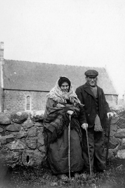 Two people in hats and scarves, standing by a drystone wall outside a small, stone church. Image reference number: SC_1105214 