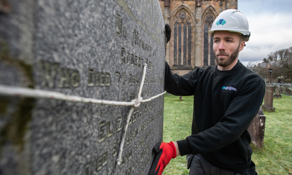 Man in white hard hat with Historic Environment Scotland logo inspects a memorial stone. The stone has string tied around it.