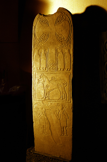 A tall, slim, upright standing stone inside a building, intricately carved with pictures and symbols