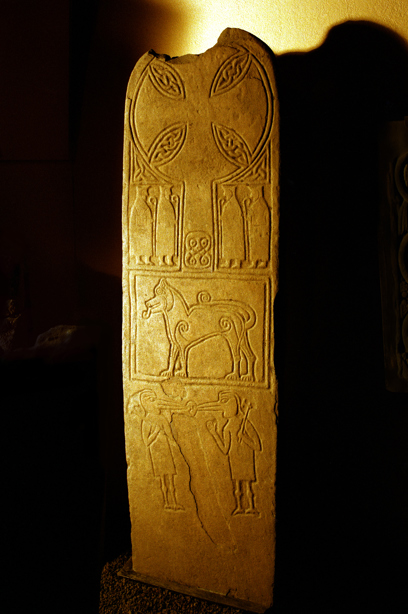 A tall, slim, upright standing stone inside a building, intricately carved with pictures and symbols