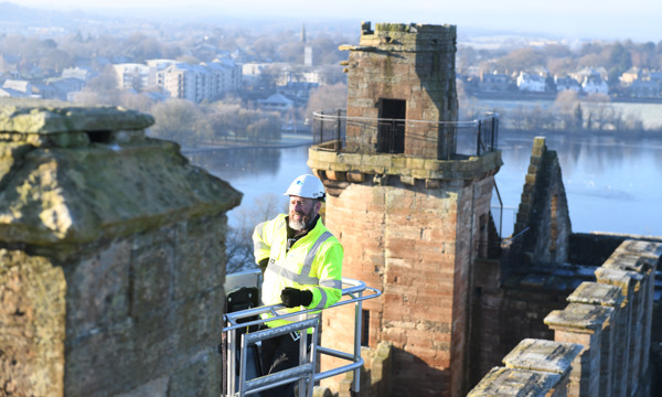 A man with a white beard, wearing a HES safety helmet and high visibility jacket stand on a high platform at the top of Linlithgow Palace. In the background we wee a lake and the local area.