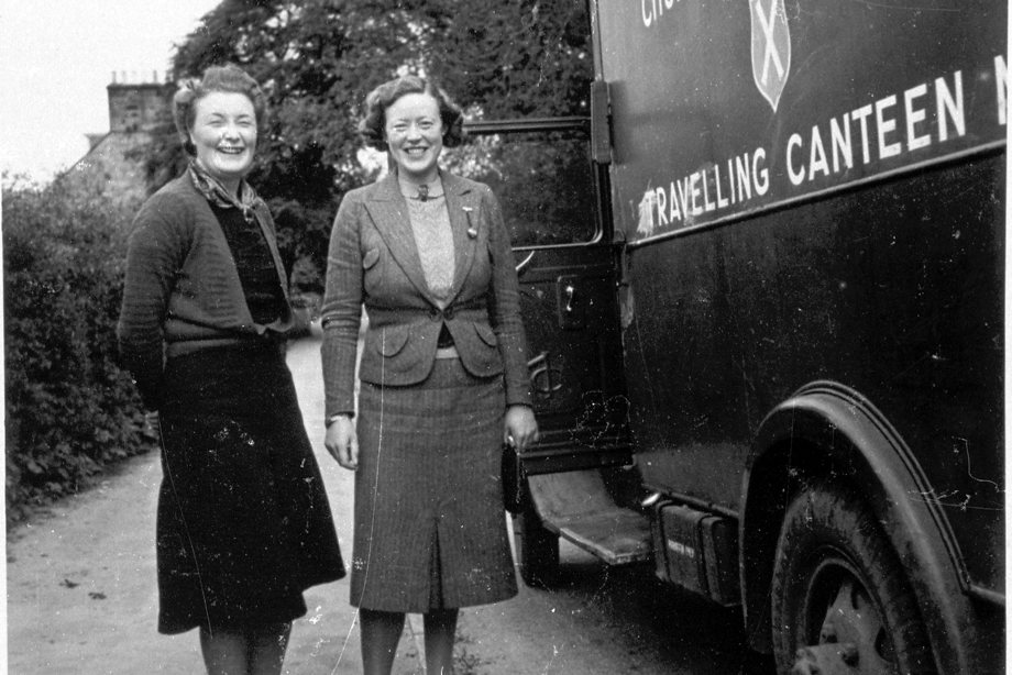 Two women dressed in skirts and blazers, standing beside a bus with 'travelling canteen' printed on the side of it.