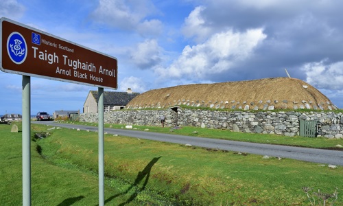 General view of the Arnol Blackhouse which is a low, stone built dwelling with a thatched roof. In front is a sign for the building in Gaelic.