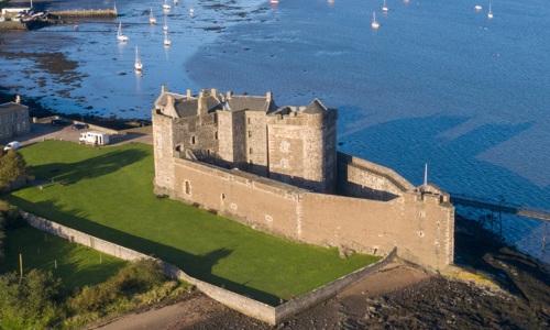 A historic castle with a large wall surrounding it, on the edge of an estuary 