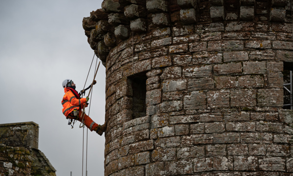 A person in PPE, including a hard hat and bright orange jumpsuit, abseils down the outer wall of Caerlaverock Castle
