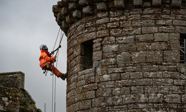 A person in PPE, including a hard hat and bright orange jumpsuit, abseils down the outer wall of Caerlaverock Castle