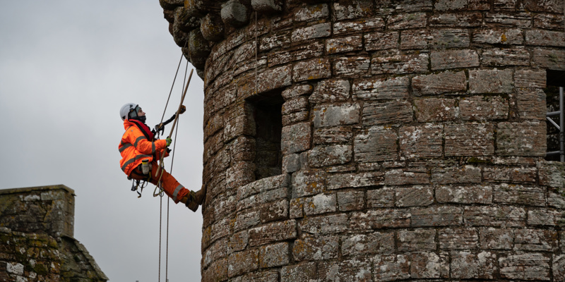 A person in PPE, including a hard hat and bright orange jumpsuit, abseils down the outer wall of Caerlaverock Castle
