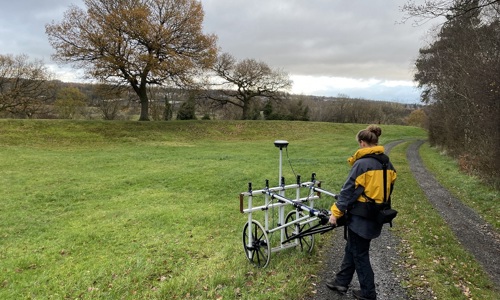 A photo of a grassy landscape and a person wheeling a piece of geophysical surveying equipment.