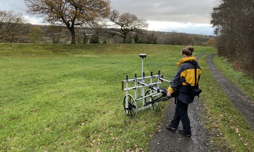 A photo of a grassy landscape and a person wheeling a piece of geophysical surveying equipment.