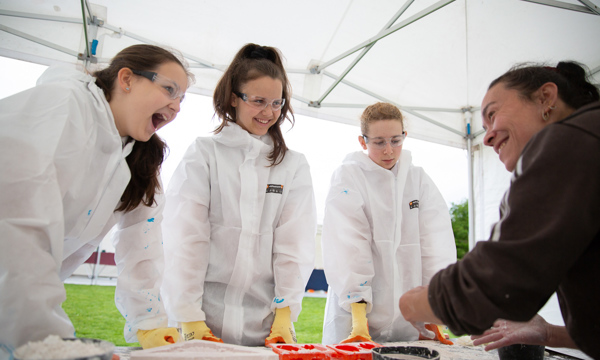 A photo of a person demonstrating traditional building skills to three young people wearing protective goggles and gloves and protective suits.