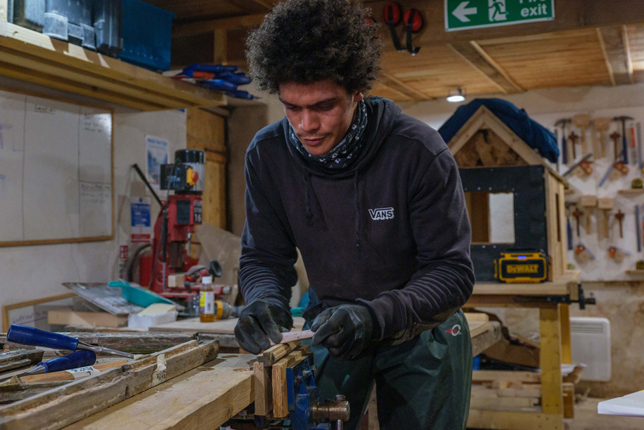 A client works within the workshop at the Ridge; the client is clothed in work gear and is sanding a piece of wood