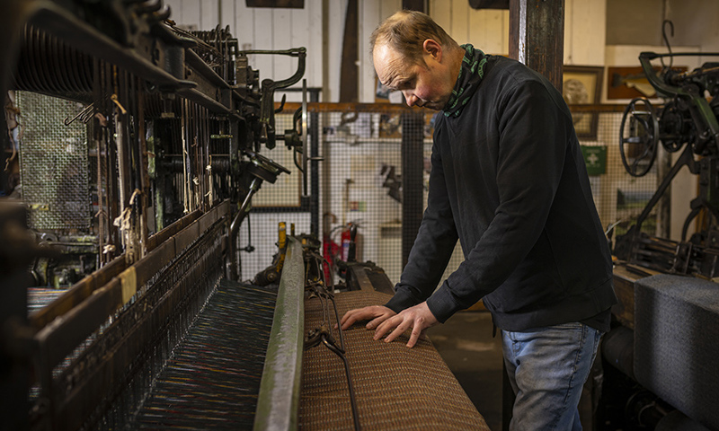 Man in jeans and a jumper works in woolen mill. Tools hang from the walls in the background.