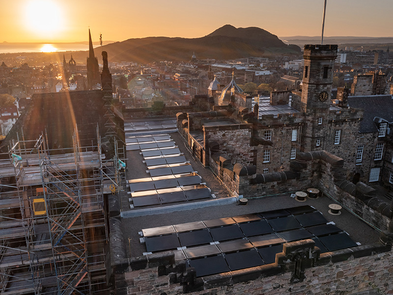 Sun rises over the city of Edinburgh and sunbeams spray across Edinburgh Castle from above, where an array of solar panels fill a flat section of the rooftop. Arthur's Seat appears in silhouette in the background with the city in between.