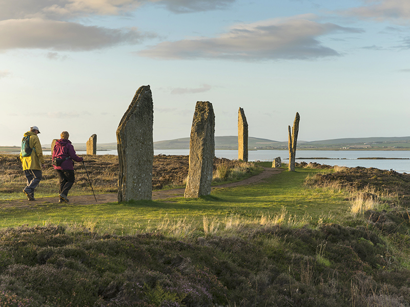 Two walkers approach standing stones along a well-trodden trail in a field. The stones stand approximately 10 foot high, twice the height of the walkers, and curve in the distance in a semi circle. The sky is blue with a smattering of clouds, in the background a large body of water water separates the field of stand stones from more land in the distance.