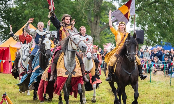 Five people dressed up in medieval clothing on horses gallop towards the camera.