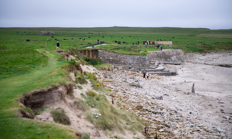 A grassy path is right on the edge of an eroding sandy bank that drops down to a beach. In the distance there is a concrete structure supporting the land above the beach where people are standing.