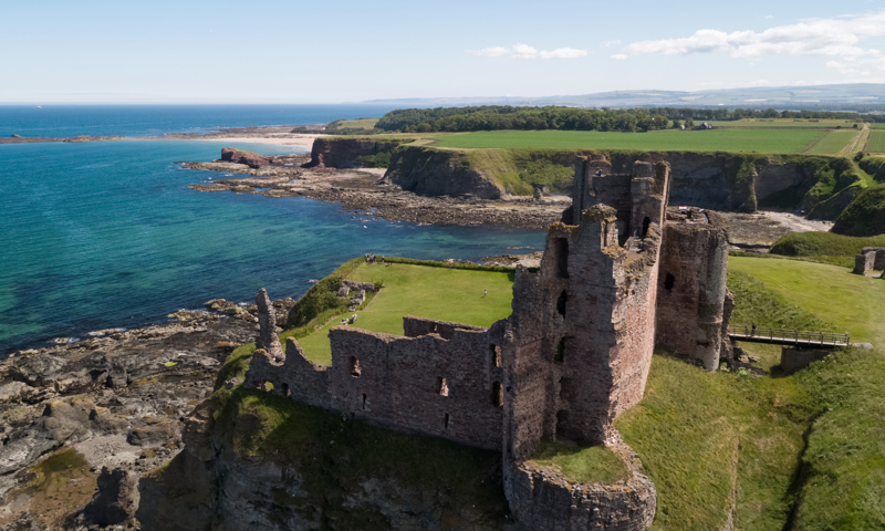Aerial view of a ruined castle with it's boundaries going right up to the cliff edge, beyond which is the sea and further coastline in the distance.