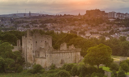 A bird's eye view of Craigmillar Castle at sunrise. You can see the castle ruin in full nestles between trees against the backdrop of the Edinburgh skyline.