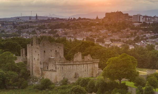A bird's eye view of Craigmillar Castle at sunrise. You can see the castle ruin in full nestles between trees against the backdrop of the Edinburgh skyline.