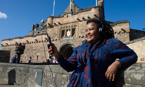 A lady on Edinburgh Castle esplanade wearing headphones and smiling looking at her mobile phone