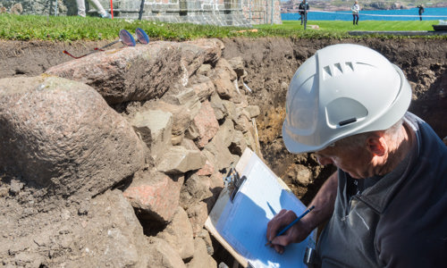 A man in a hard hat standing in a hole in a trench in the ground with a clipboard