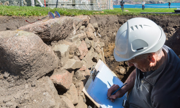 A man in a hard hat standing in a hole in a trench in the ground with a clipboard