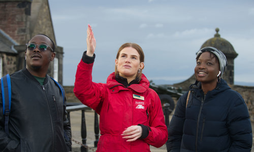 A member of staff at Edinburgh Castle wearing a branded jacket pointing out something to two visitors 
