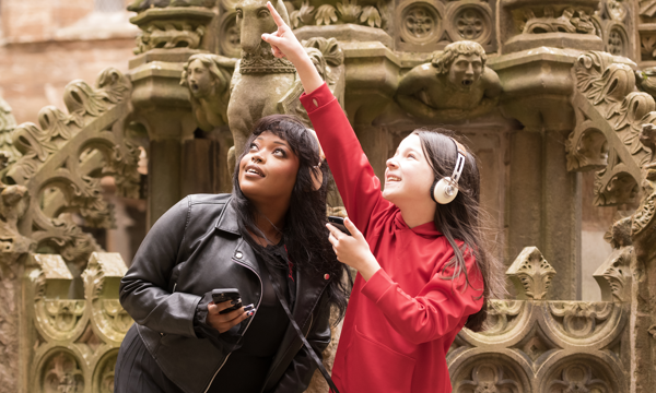 A young woman and a girl stand in front of ornate stone carvings. The girl wears a red top and head phones and she points at something out of shot. The young woman is dressed in a black leather jacket and also wears headphones. She looks in the direction the girl is pointing.