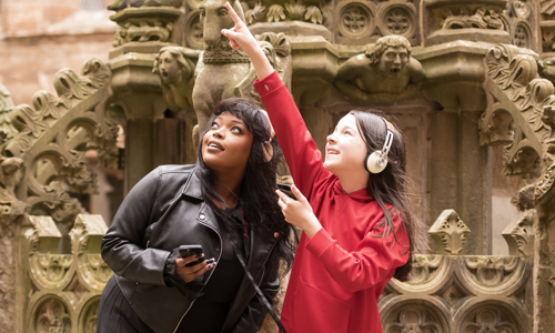 A young woman and a girl stand in front of ornate stone carvings. The girl wears a red top and head phones and she points at something out of shot. The young woman is dressed in a black leather jacket and also wears headphones. She looks in the direction the girl is pointing.