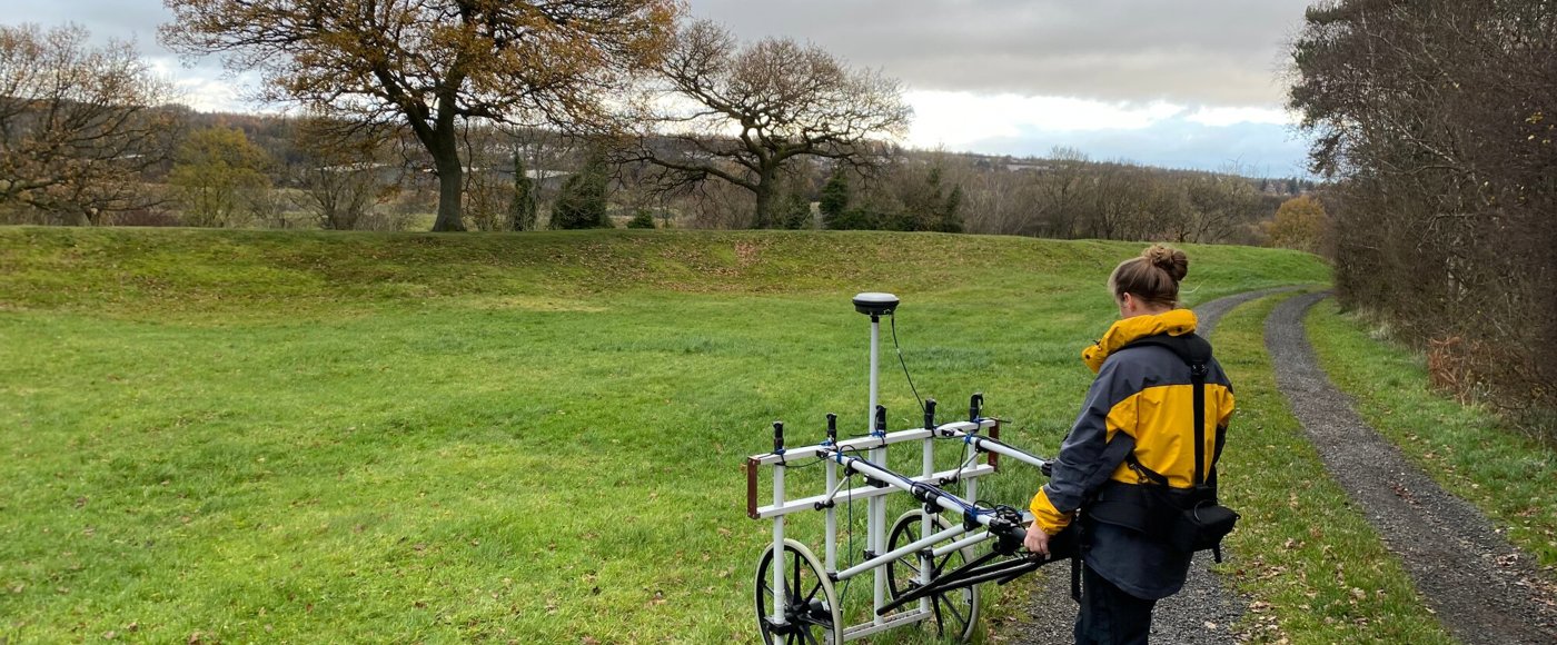 Image shows a person wearing outdoor clothing, standing behind and pushing a piece of scientific equipment across a grassy area with trees behind. The equipment is a large white lattice, about the size of a door on it side, attached to wheels.