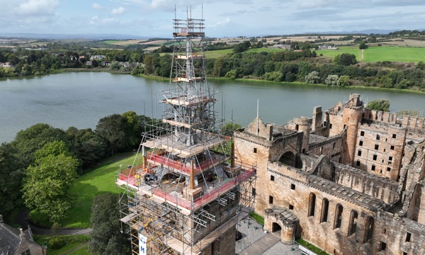 Church spire shown wrapped in scaffolding with Linlithgow Palace behind. The background shows Linlithgow Loch surrounded by trees and fields.