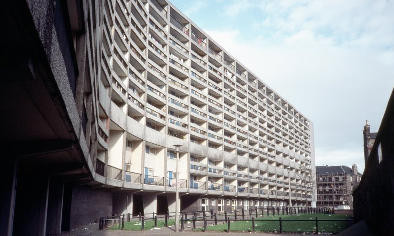 Black and white image of a block of flats, with scattered clouds in the sky. The flats are made of concrete and brutalist in style, with many small windows and balconies. The block has a bend along the centre hence the nickname 