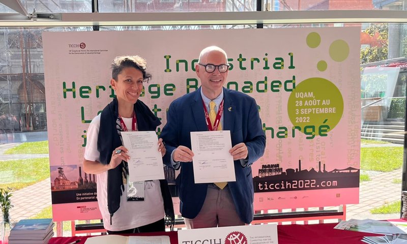 Two people standing behind a red desk outdoors in the sunshine. They are both holding a single sheet of white paper and smiling at the camera.