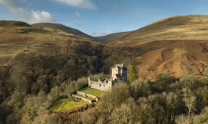 An aerial view of a large grey castle and its grounds surrounded by trees. There are open hills in the background.