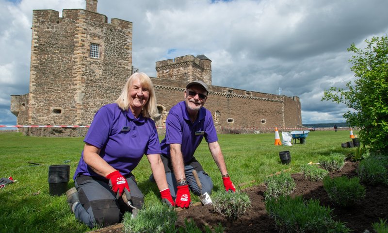 Two people on their knees are tending to a flower bed. They are smiling and holding gardening tools. Behind them is a large semi ruined castle.