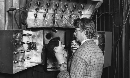 Black and white photograph of a person seated behind a microphone and a bank of light bulbs. They are holding a ventriloquist
